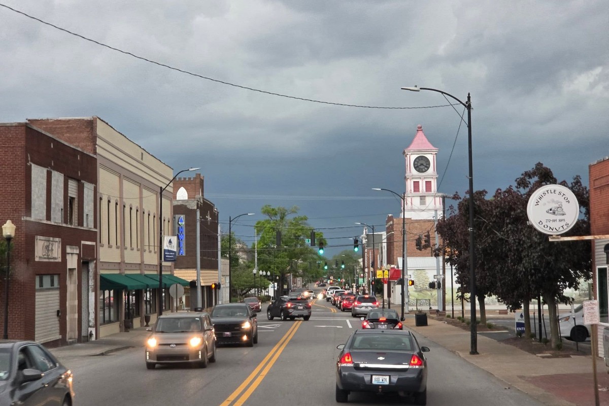 Busy main street with brick storefronts and moving cars, a pink-roofed clock tower visible in the distance under a cloudy sky.