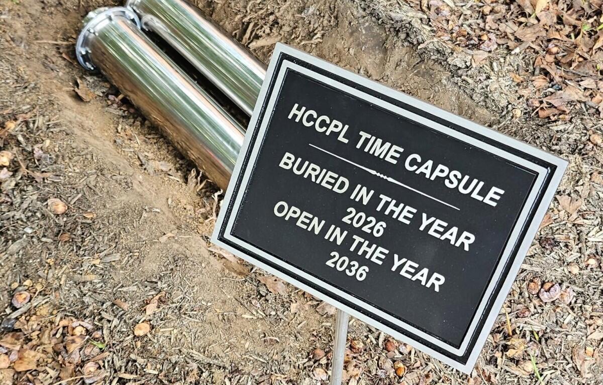 Metal time capsule and sign: 'HCCPL Time Capsule' buried in 2026, to be opened in 2036, set in dirt with fallen leaves.