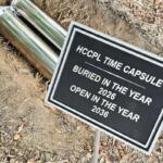 Metal time capsule and sign: 'HCCPL Time Capsule' buried in 2026, to be opened in 2036, set in dirt with fallen leaves.