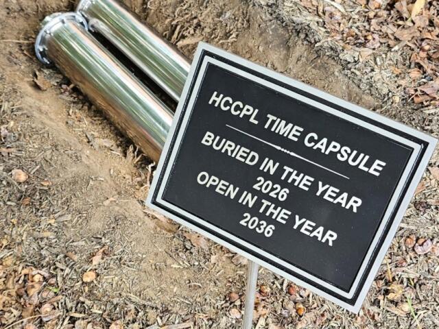 Metal time capsule and sign: 'HCCPL Time Capsule' buried in 2026, to be opened in 2036, set in dirt with fallen leaves.