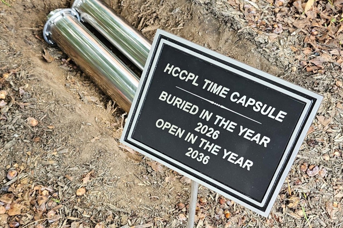Metal time capsule and sign: 'HCCPL Time Capsule' buried in 2026, to be opened in 2036, set in dirt with fallen leaves.