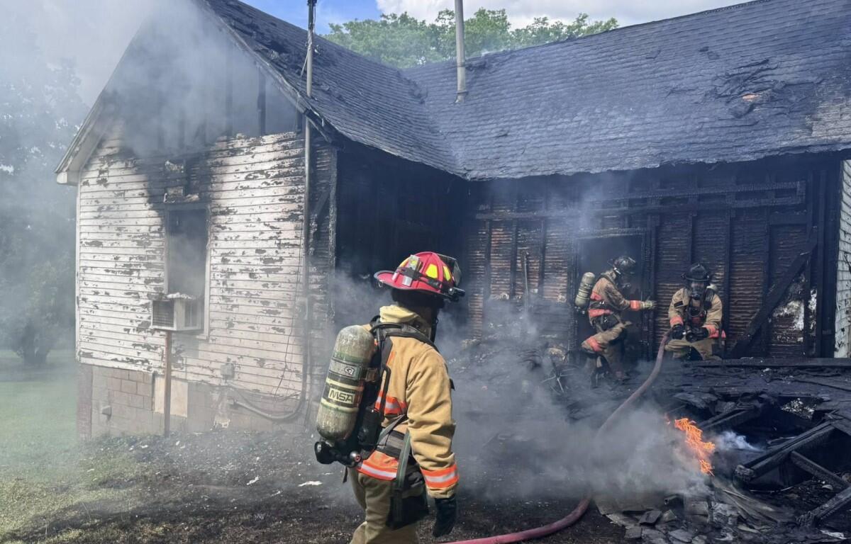 Firefighters in full turnout gear extinguish a smoke-filled, charred house fire at a damaged home exterior.