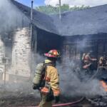 Firefighters in full turnout gear extinguish a smoke-filled, charred house fire at a damaged home exterior.