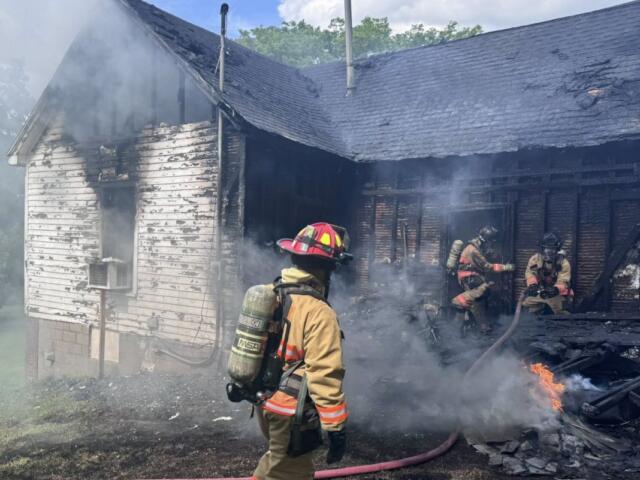 Firefighters in full turnout gear extinguish a smoke-filled, charred house fire at a damaged home exterior.