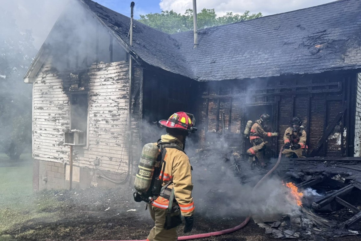 Firefighters in full turnout gear extinguish a smoke-filled, charred house fire at a damaged home exterior.