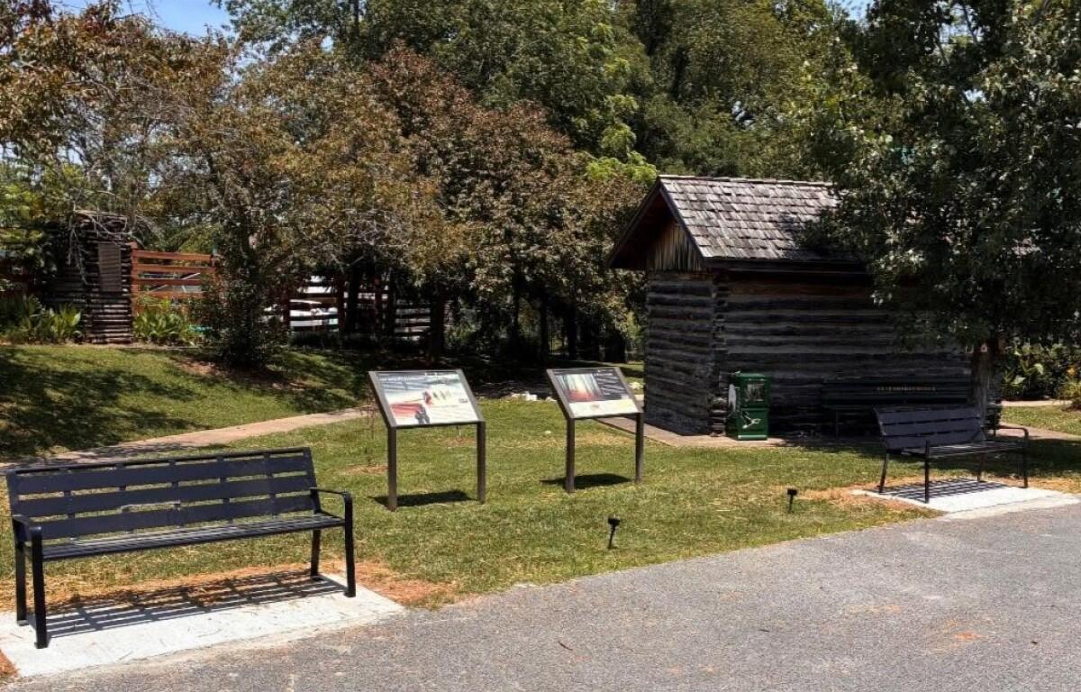 Park setting with two black metal benches, two informational display panels, and a small log cabin among trees.