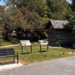 Park setting with two black metal benches, two informational display panels, and a small log cabin among trees.