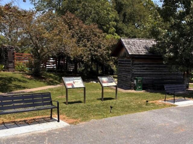 Park setting with two black metal benches, two informational display panels, and a small log cabin among trees.