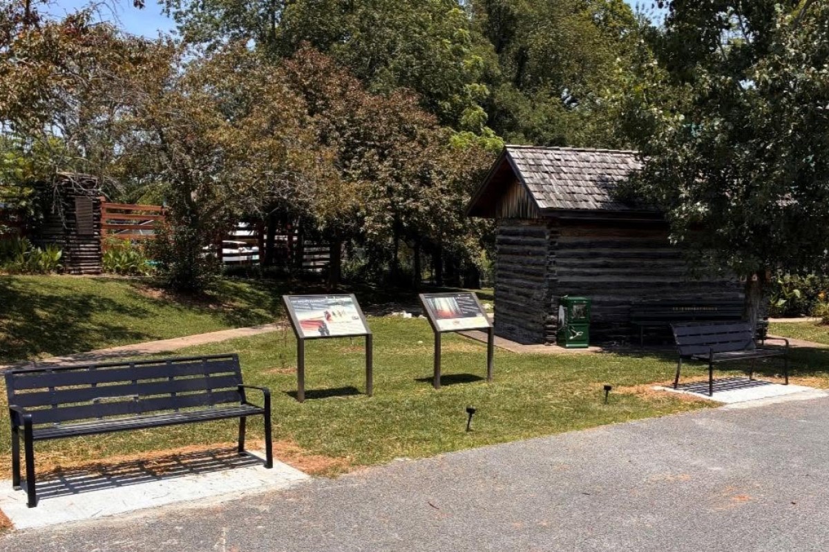 Park setting with two black metal benches, two informational display panels, and a small log cabin among trees.