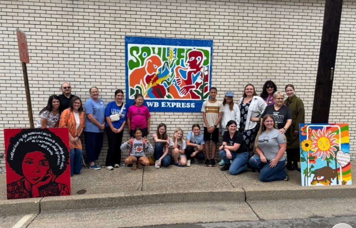 Group of adults and kids posing with colorful paintings on a sidewalk in front of a brick wall and a blue WB EXPRESS mural sign.