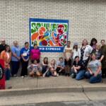 Group of adults and kids posing with colorful paintings on a sidewalk in front of a brick wall and a blue WB EXPRESS mural sign.