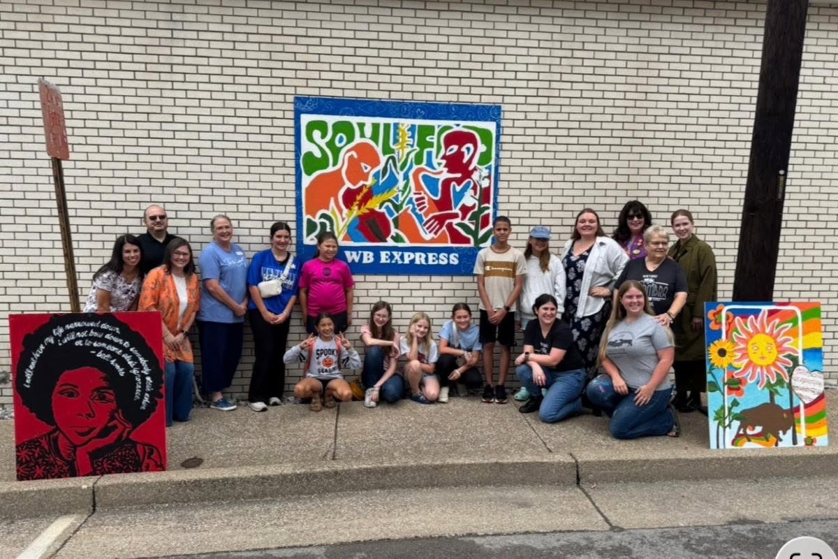 Group of adults and kids posing with colorful paintings on a sidewalk in front of a brick wall and a blue WB EXPRESS mural sign.