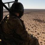 Soldier in camouflage gear and helmet sits at the edge of a helicopter, looking out over a barren desert with sparse shrubs.