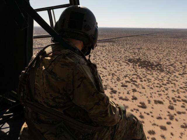 Soldier in camouflage gear and helmet sits at the edge of a helicopter, looking out over a barren desert with sparse shrubs.