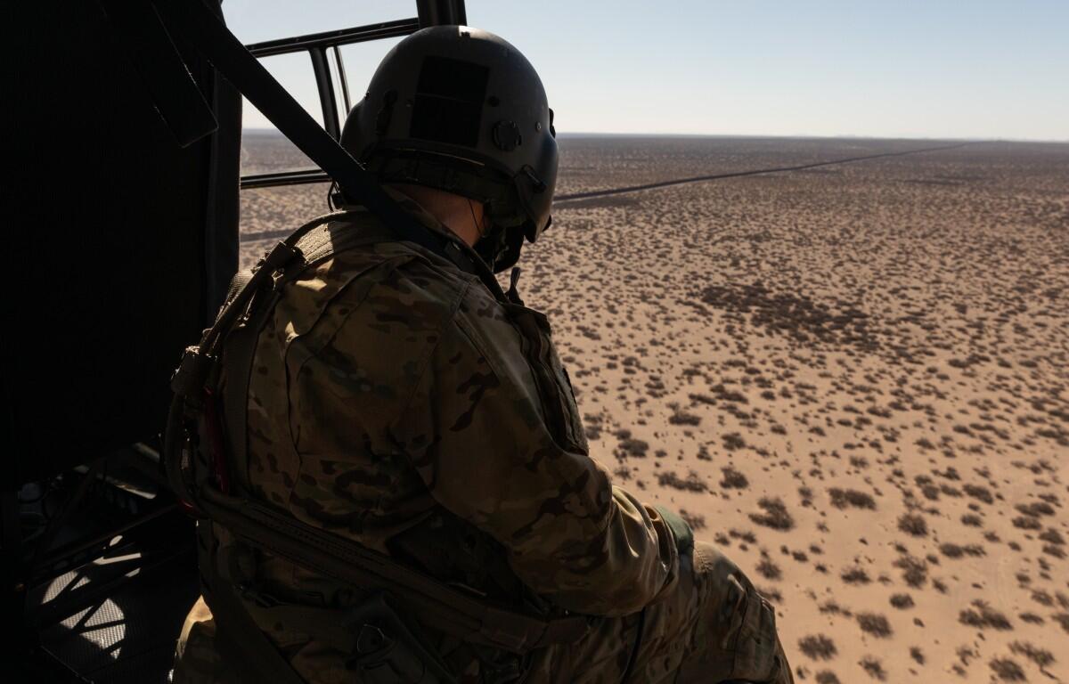 Soldier in camouflage gear and helmet sits at the edge of a helicopter, looking out over a barren desert with sparse shrubs.