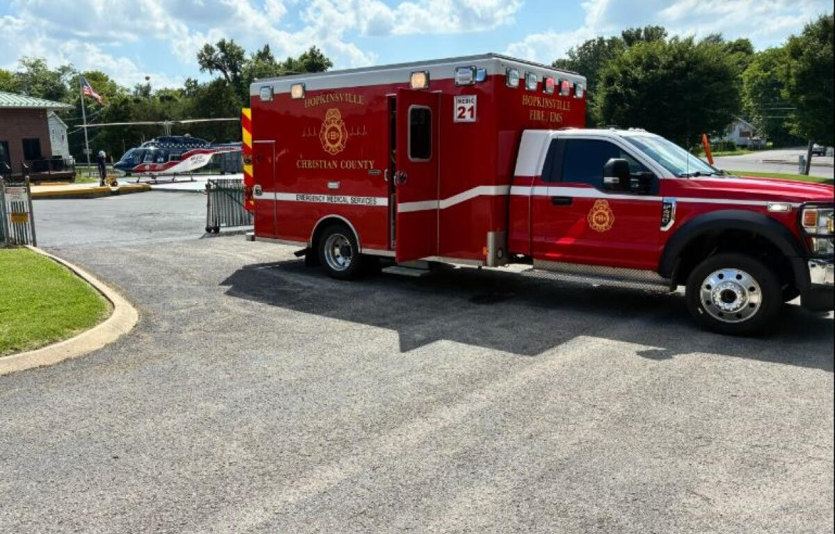 Red emergency medical services ambulance parked in a lot beside a building, with a small helicopter visible in the background under a blue sky.