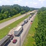 Traffic jam on a highway: multiple trucks and cars lined up in the right lanes near a construction area with orange cones and an overhead sign gantry.