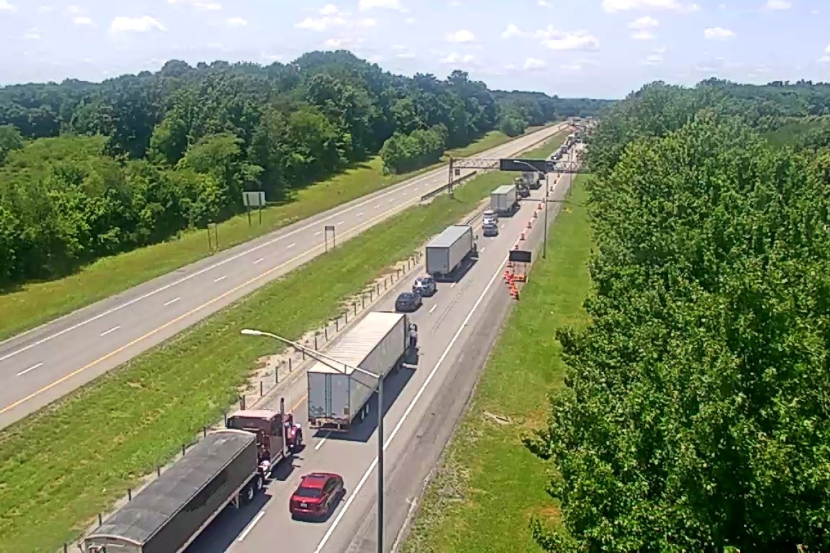 Traffic jam on a highway: multiple trucks and cars lined up in the right lanes near a construction area with orange cones and an overhead sign gantry.