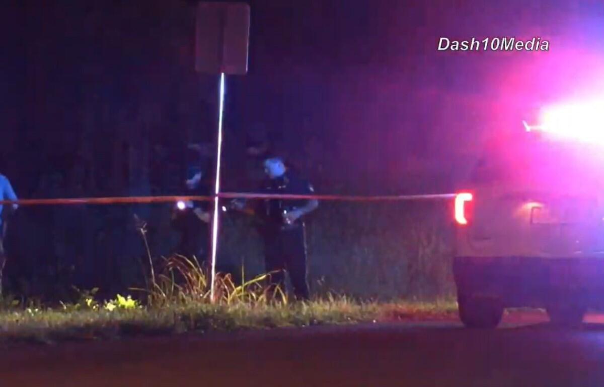 Nighttime scene with police car flashing red/blue lights, orange caution tape across a roadside and two officers behind it near a signpost.