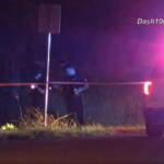 Nighttime scene with police car flashing red/blue lights, orange caution tape across a roadside and two officers behind it near a signpost.