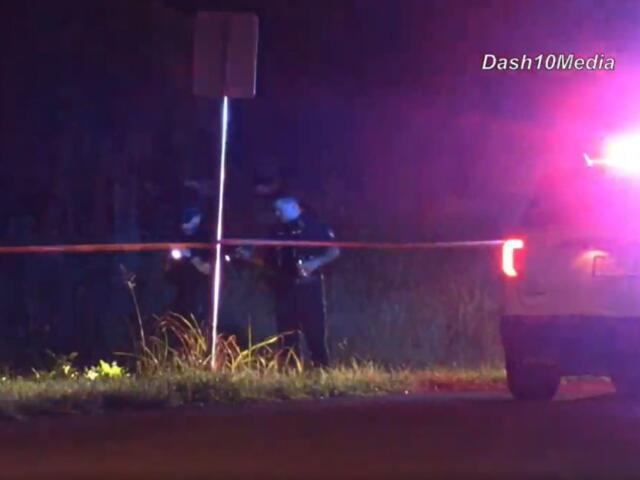 Nighttime scene with police car flashing red/blue lights, orange caution tape across a roadside and two officers behind it near a signpost.