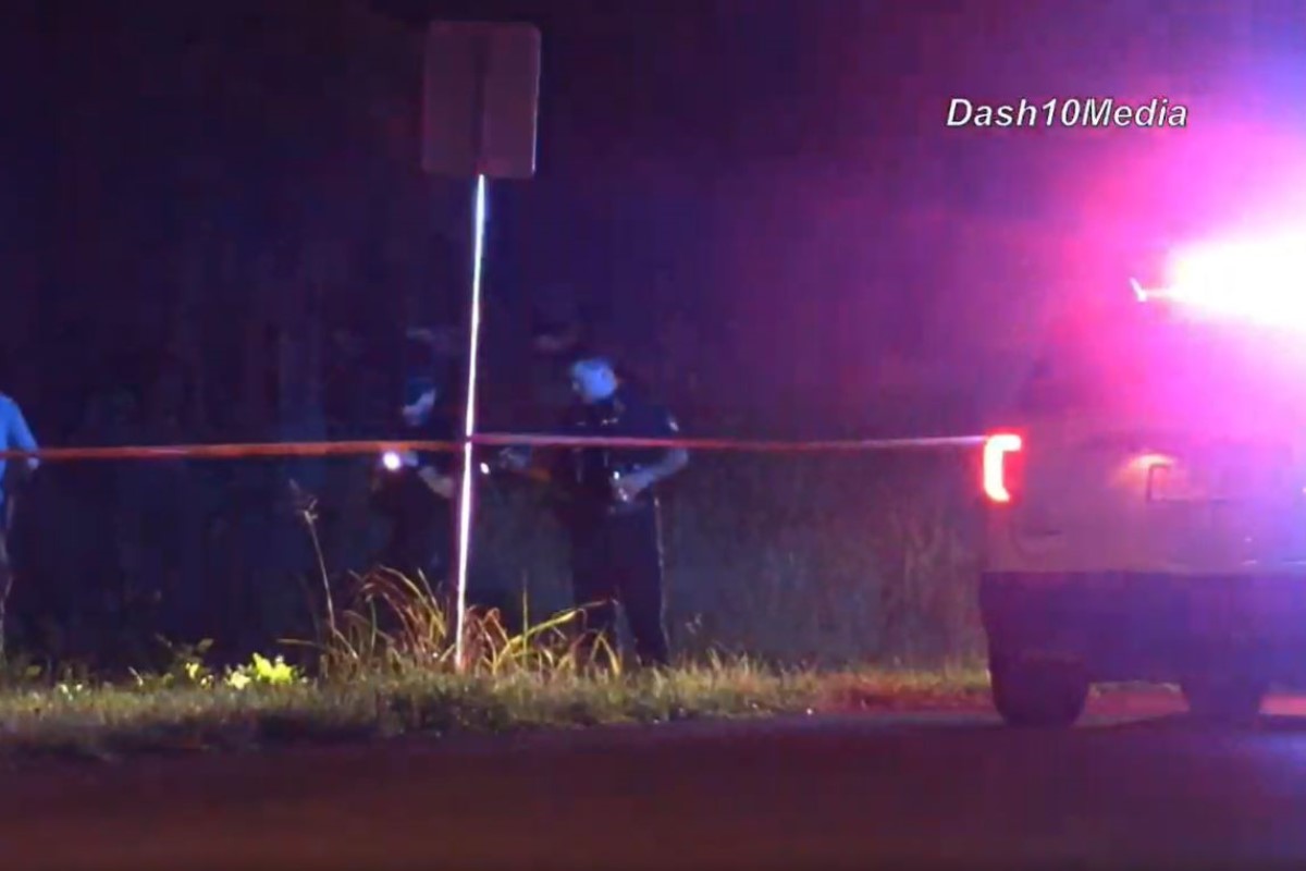 Nighttime scene with police car flashing red/blue lights, orange caution tape across a roadside and two officers behind it near a signpost.