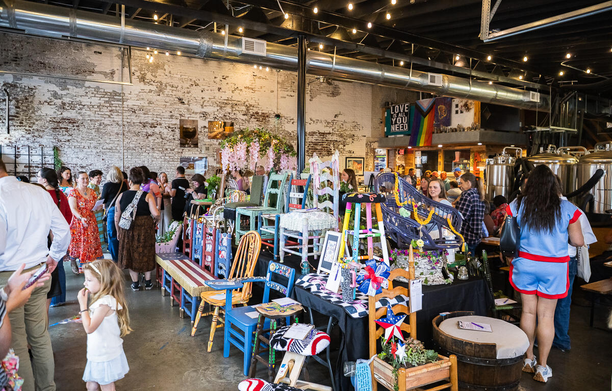 Crowd browsing handmade furniture and crafts on display tables in a rustic industrial market space with string lights above. A stall of colorful painted chairs and decorative items set up on tables as shoppers explore in a brick-walled venue. Indoor craft fair scene with vendors selling painted chairs, plants, and handmade decor under warm string lights. People mingle around a vendor table of colorful wooden chairs and ornaments in an industrial-style market. A young girl in a white dress stands near the display while others browse the market stalls.