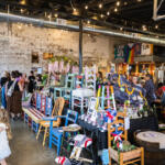 Crowd browsing handmade furniture and crafts on display tables in a rustic industrial market space with string lights above. A stall of colorful painted chairs and decorative items set up on tables as shoppers explore in a brick-walled venue. Indoor craft fair scene with vendors selling painted chairs, plants, and handmade decor under warm string lights. People mingle around a vendor table of colorful wooden chairs and ornaments in an industrial-style market. A young girl in a white dress stands near the display while others browse the market stalls.