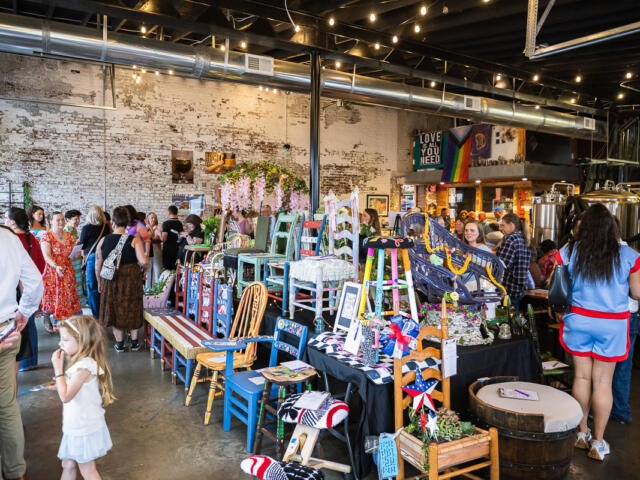 Crowd browsing handmade furniture and crafts on display tables in a rustic industrial market space with string lights above. A stall of colorful painted chairs and decorative items set up on tables as shoppers explore in a brick-walled venue. Indoor craft fair scene with vendors selling painted chairs, plants, and handmade decor under warm string lights. People mingle around a vendor table of colorful wooden chairs and ornaments in an industrial-style market. A young girl in a white dress stands near the display while others browse the market stalls.