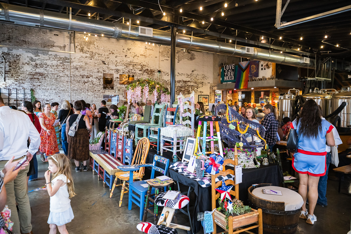 Crowd browsing handmade furniture and crafts on display tables in a rustic industrial market space with string lights above. A stall of colorful painted chairs and decorative items set up on tables as shoppers explore in a brick-walled venue. Indoor craft fair scene with vendors selling painted chairs, plants, and handmade decor under warm string lights. People mingle around a vendor table of colorful wooden chairs and ornaments in an industrial-style market. A young girl in a white dress stands near the display while others browse the market stalls.