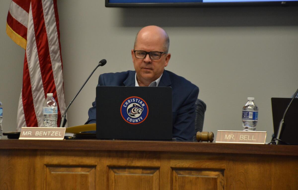 Man in a suit and glasses sits at a panel desk with a microphone, gavel, water bottle, and a laptop displaying a Christian County seal; nameplate reads 'MR. BENTZEL'.