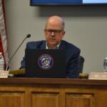 Man in a suit and glasses sits at a panel desk with a microphone, gavel, water bottle, and a laptop displaying a Christian County seal; nameplate reads 'MR. BENTZEL'.