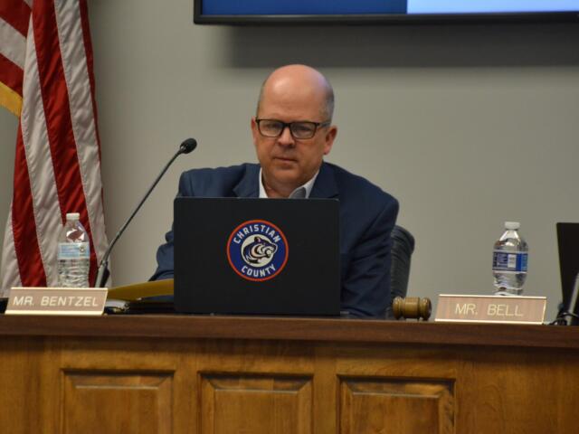 Man in a suit and glasses sits at a panel desk with a microphone, gavel, water bottle, and a laptop displaying a Christian County seal; nameplate reads 'MR. BENTZEL'.