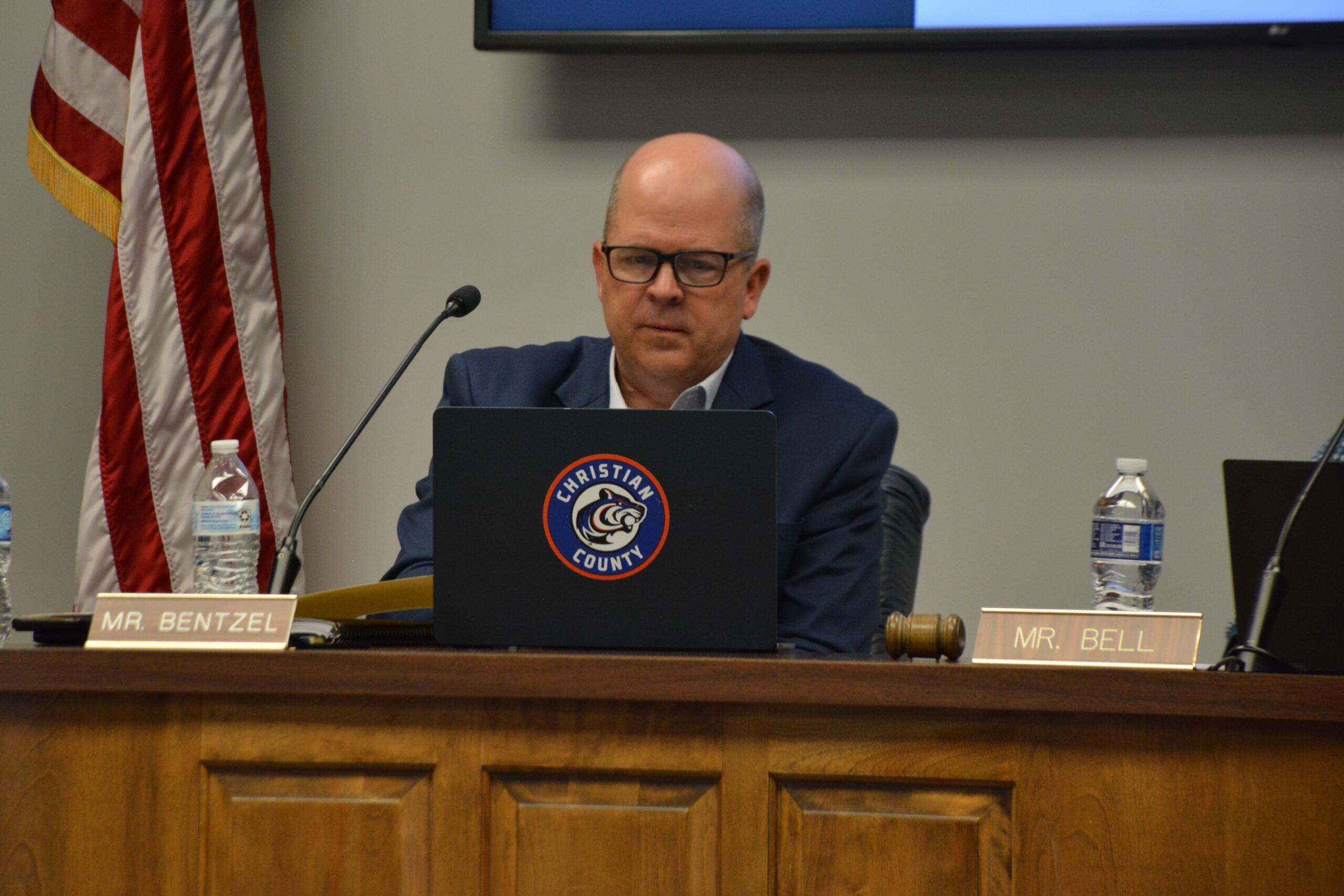 Man in a suit and glasses sits at a panel desk with a microphone, gavel, water bottle, and a laptop displaying a Christian County seal; nameplate reads 'MR. BENTZEL'.