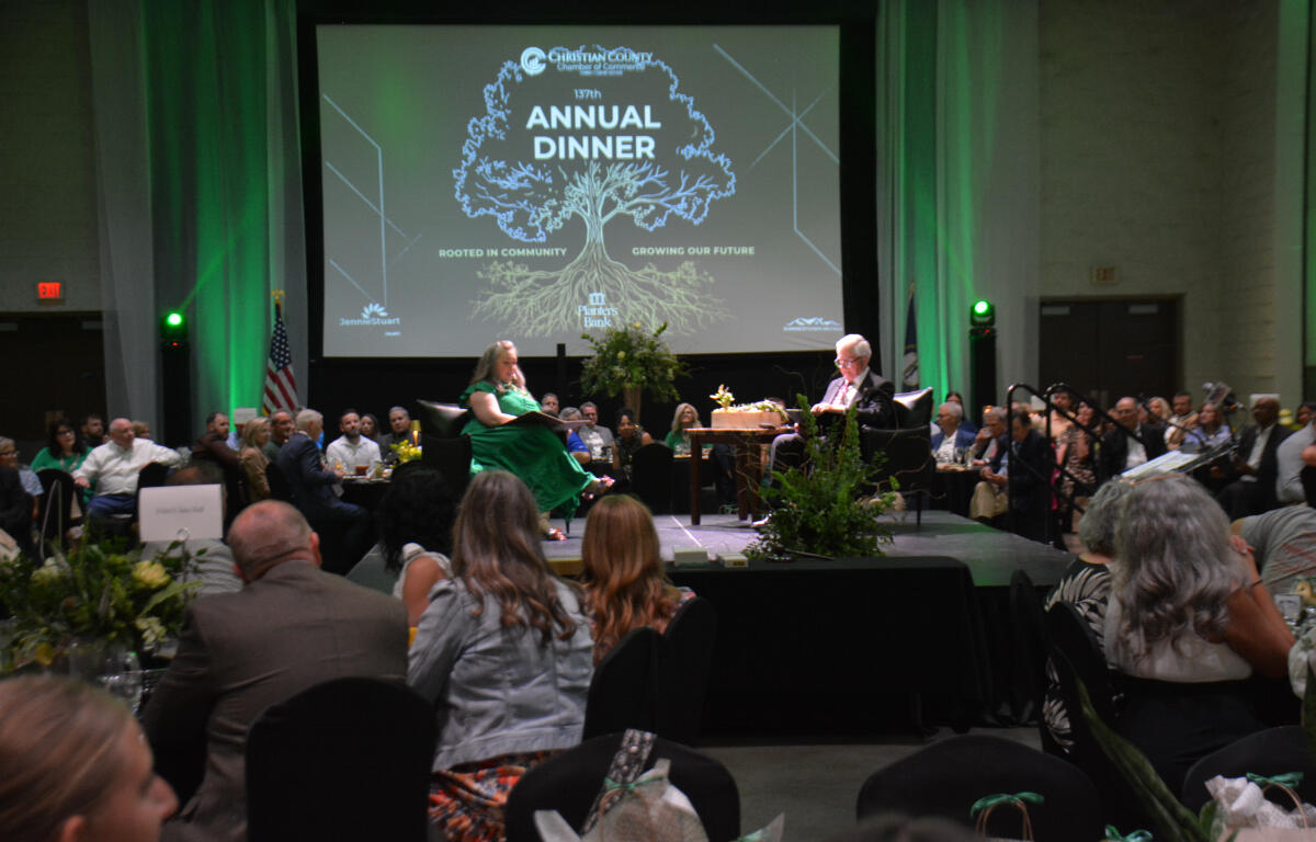 Audience seated at round tables in a large hall, two people on a stage in front of a screen that reads 'Annual Dinner' with a tree graphic.