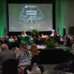 Audience seated at round tables in a large hall, two people on a stage in front of a screen that reads 'Annual Dinner' with a tree graphic.