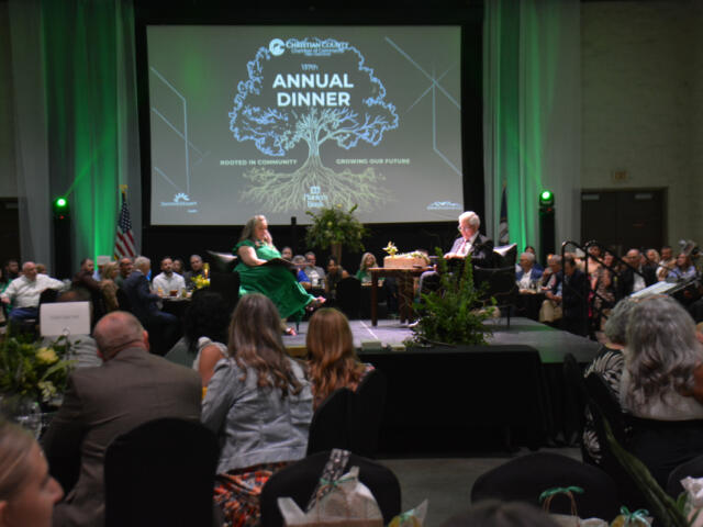 Audience seated at round tables in a large hall, two people on a stage in front of a screen that reads 'Annual Dinner' with a tree graphic.