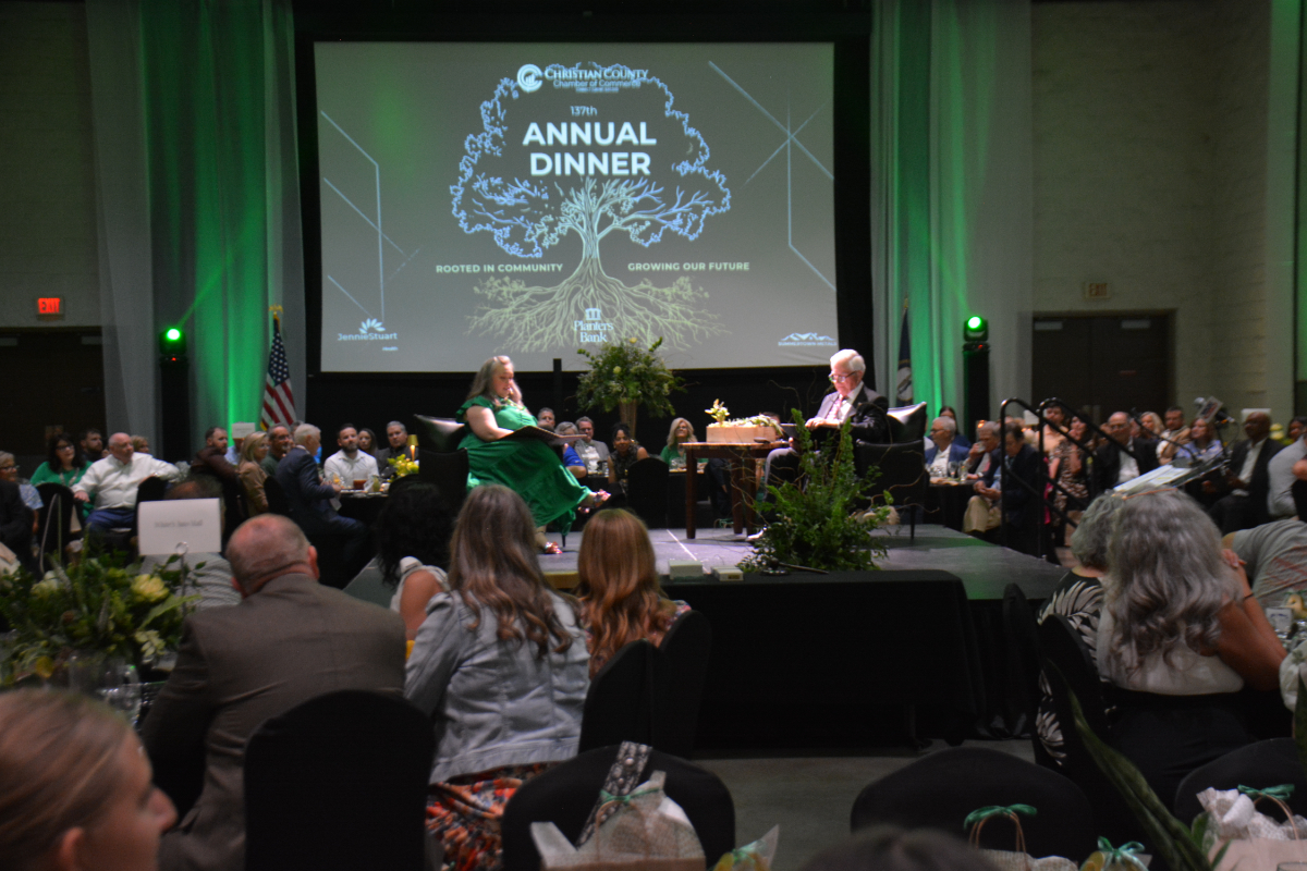 Audience seated at round tables in a large hall, two people on a stage in front of a screen that reads 'Annual Dinner' with a tree graphic.
