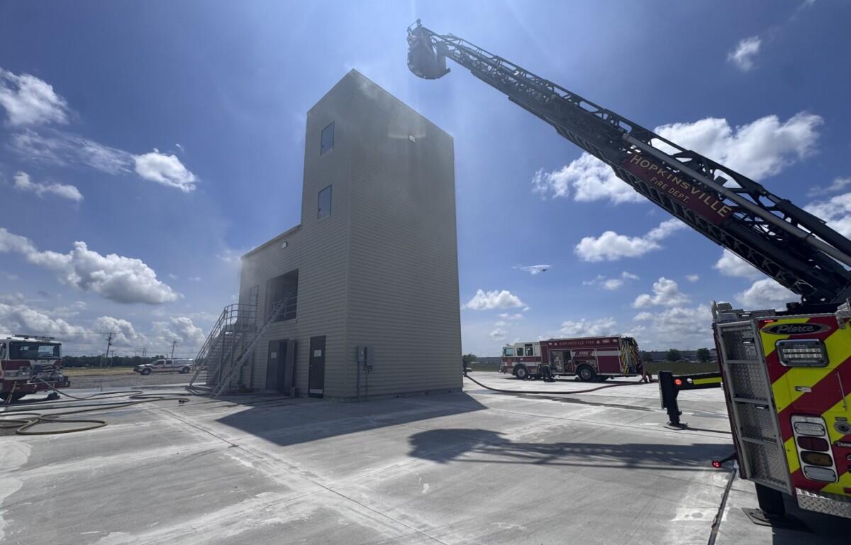 Ladder truck extending its long boom toward a tall beige training tower on a sunny day at a fire facility.