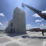 Ladder truck extending its long boom toward a tall beige training tower on a sunny day at a fire facility.