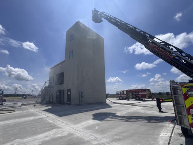 Ladder truck extending its long boom toward a tall beige training tower on a sunny day at a fire facility.