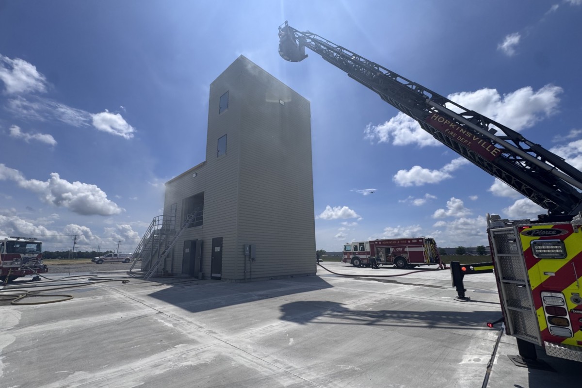 Ladder truck extending its long boom toward a tall beige training tower on a sunny day at a fire facility.