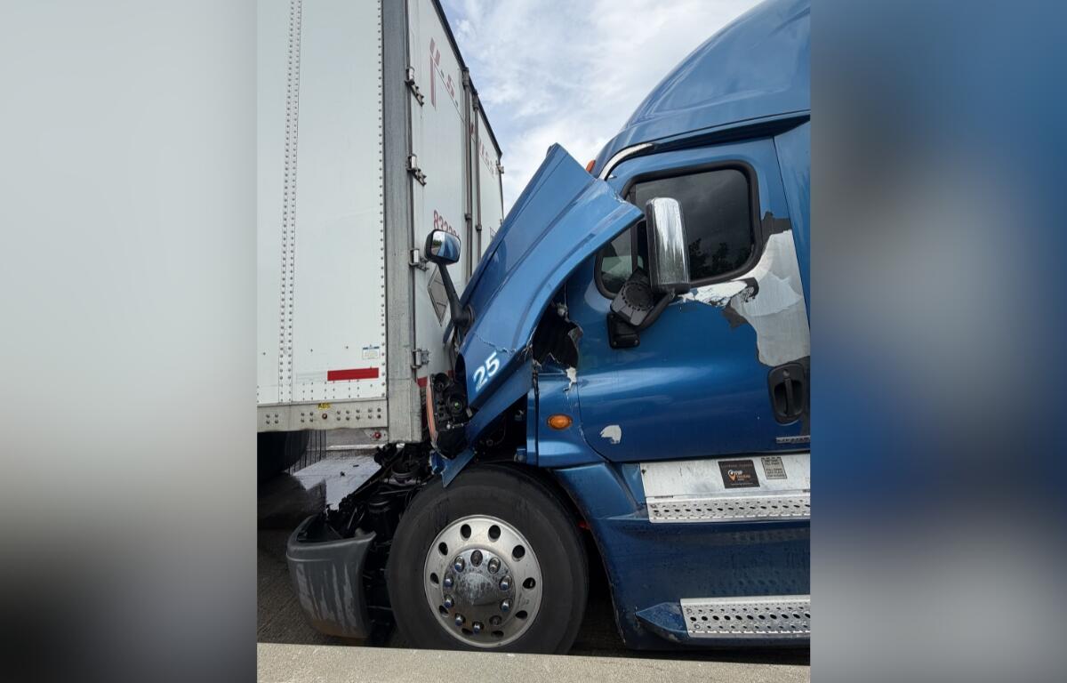 Blue semi-truck crashed into a white trailer, with the front cab severely damaged and wedged against the trailer.