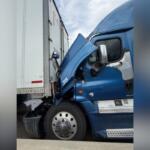Blue semi-truck crashed into a white trailer, with the front cab severely damaged and wedged against the trailer.