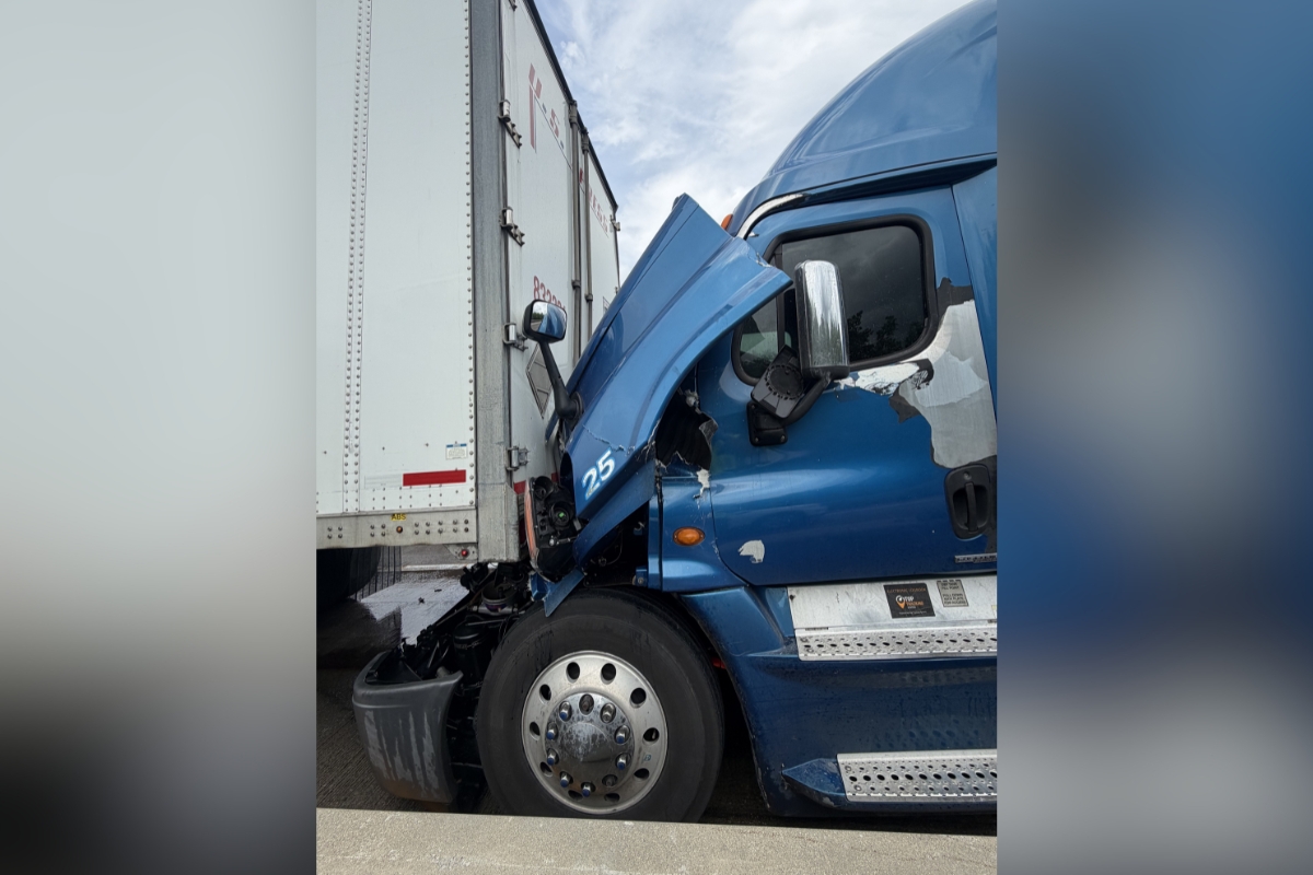 Blue semi-truck crashed into a white trailer, with the front cab severely damaged and wedged against the trailer.