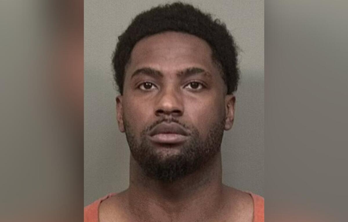 Mugshot of a man with short curly hair and facial hair wearing an orange jail jumpsuit against a grey backdrop.