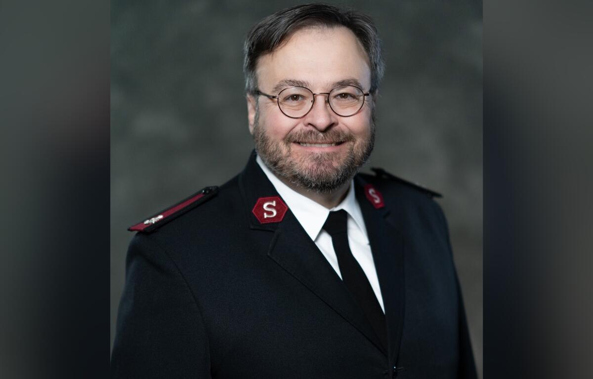 Portrait of a man in a dark formal uniform with red shoulder patches and round glasses, smiling at the camera.