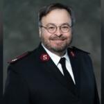Portrait of a man in a dark formal uniform with red shoulder patches and round glasses, smiling at the camera.