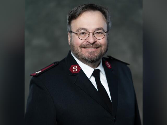 Portrait of a man in a dark formal uniform with red shoulder patches and round glasses, smiling at the camera.
