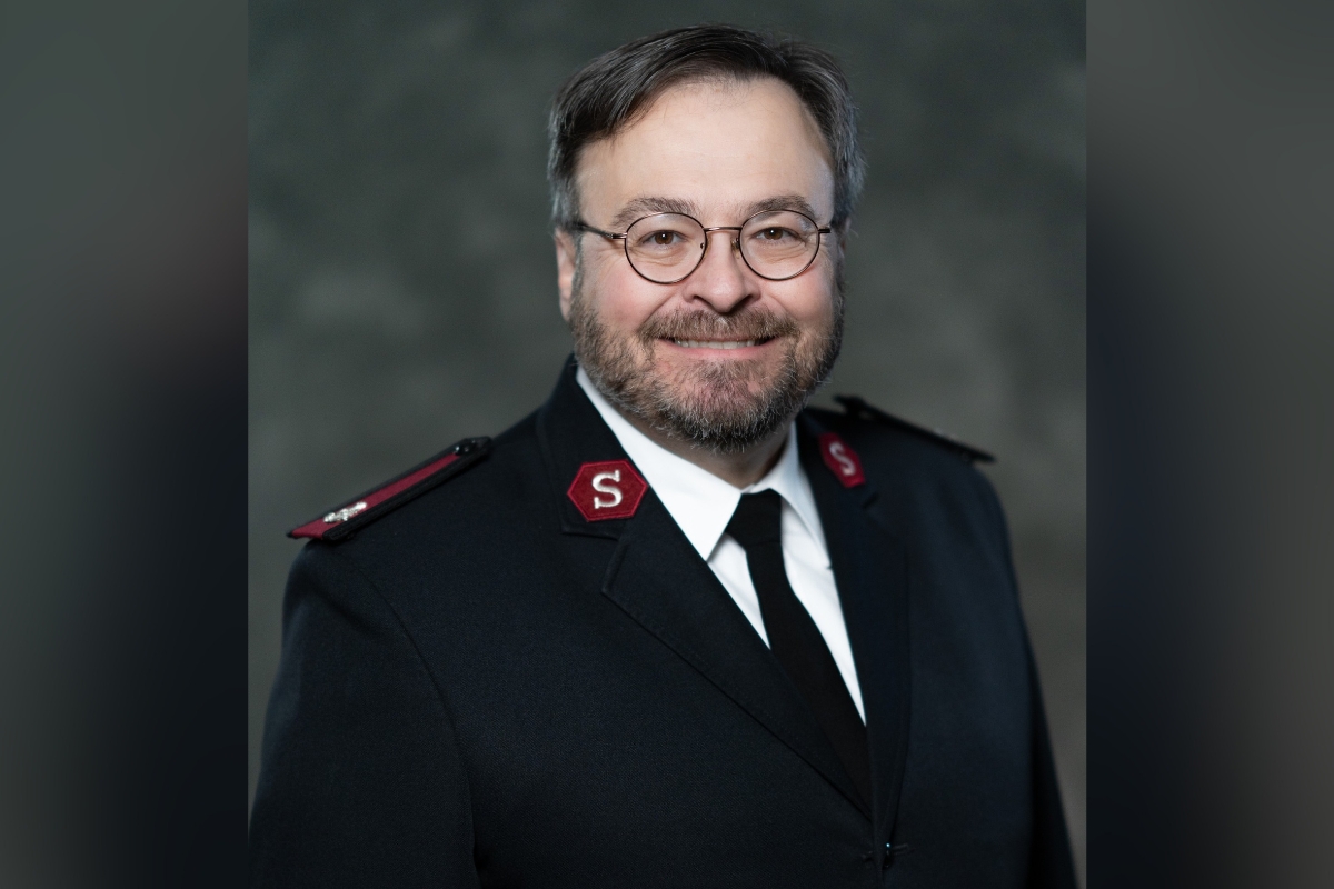 Portrait of a man in a dark formal uniform with red shoulder patches and round glasses, smiling at the camera.